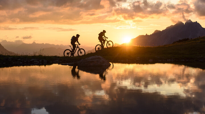 Zwei Personen fahren im Sonnenuntergang mit ihren Mountainbikes vor einer Bergkulisse. | © DAV/Wolfgang Ehn